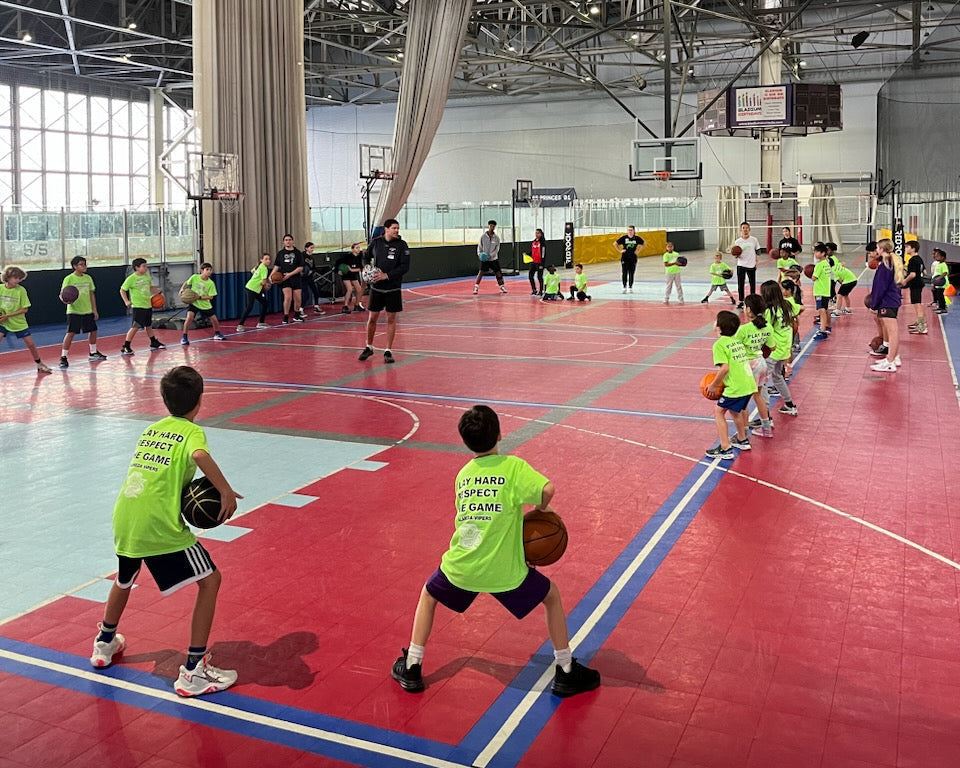Youth athletes wearing Neon Green Tees, standing on a court holding basketballs looking at the coach during Max Skills Basketball Camp in Alameda, CA.